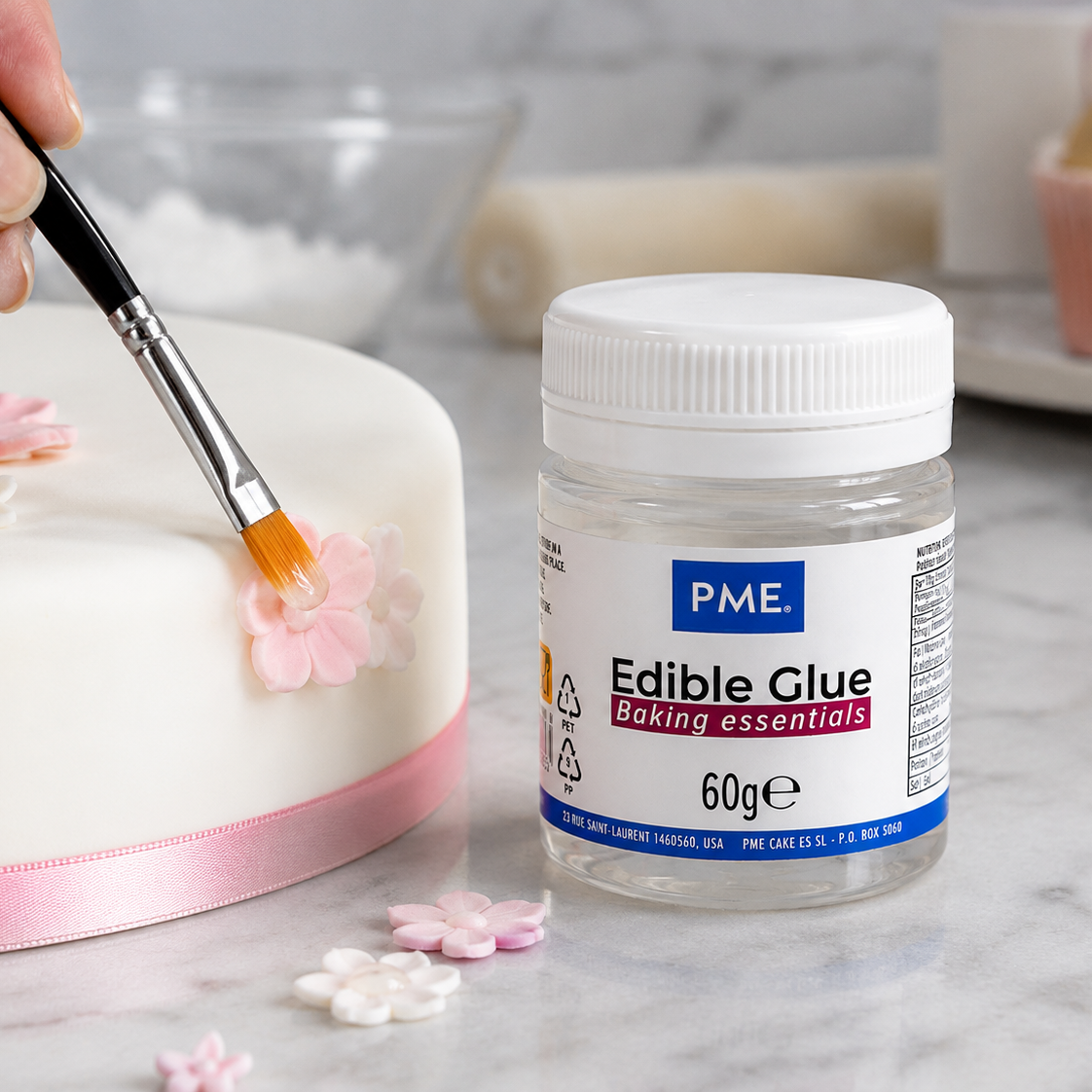 A jar of PME Edible Glue baking essentials sits on a marble countertop next to a white cake decorated with pink and white sugar flowers, with a hand holding a paintbrush applying edible glue.