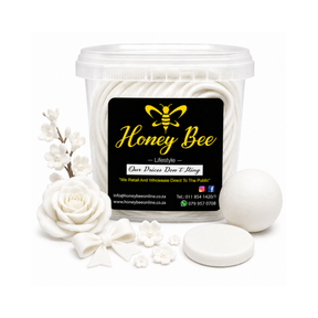 A clear plastic tub of Honey Bee white beeswax melts with a black and yellow label, shown on a white background with decorative white wax flowers, a rose, a bow, a round wax melt, and a lid.