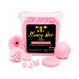 A clear plastic tub filled with pink rolled wax melts, shown with a black front label reading Honey Bee. Surrounding the tub are pink decorative flowers, a pink beeswax melt ball, a round lid, and small pink wax flower and butterfly shapes on a white background.