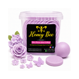 A clear plastic tub filled with lavender-colored wax melts, shown with a black front label reading Honey Bee Lifestyle. Surrounding the tub are lavender wax flower and butterfly ornaments, a lavender wax ball, and a round lavender wax disc, all on a white background.