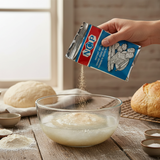 Person pouring flour from a can into a bowl with bread and other ingredients on a wooden table.