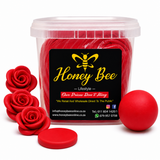 A clear plastic tub filled with bright red wax melts, featuring a black label with a yellow bee logo and the brand name 'Honey Bee'. The tub is styled with three red wax rose petals on the left, a single red wax ball on the right, and a small round red lid piece in front, all on a clean white background.