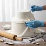 Person decorating a cake with white fondant on a table with baking tools.