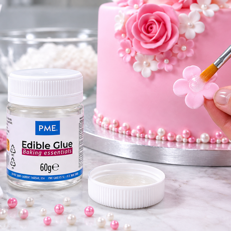 A jar of PME Edible Glue baking essentials sits on a marble surface next to a pink decorated cake, with a hand holding a paintbrush applying edible flower decorations.