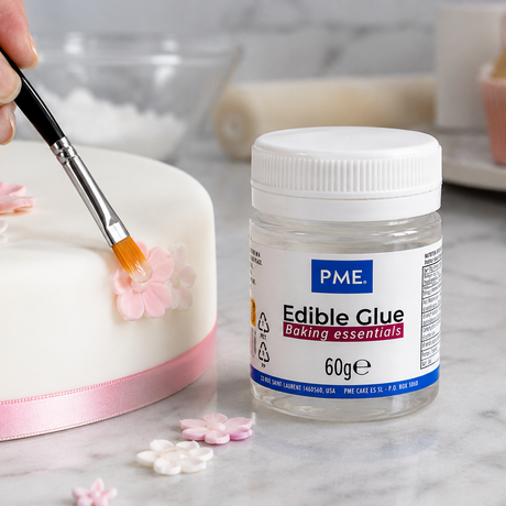 A jar of PME Edible Glue baking essentials sits on a marble countertop next to a white cake decorated with pink and white sugar flowers, with a hand holding a paintbrush applying edible glue.