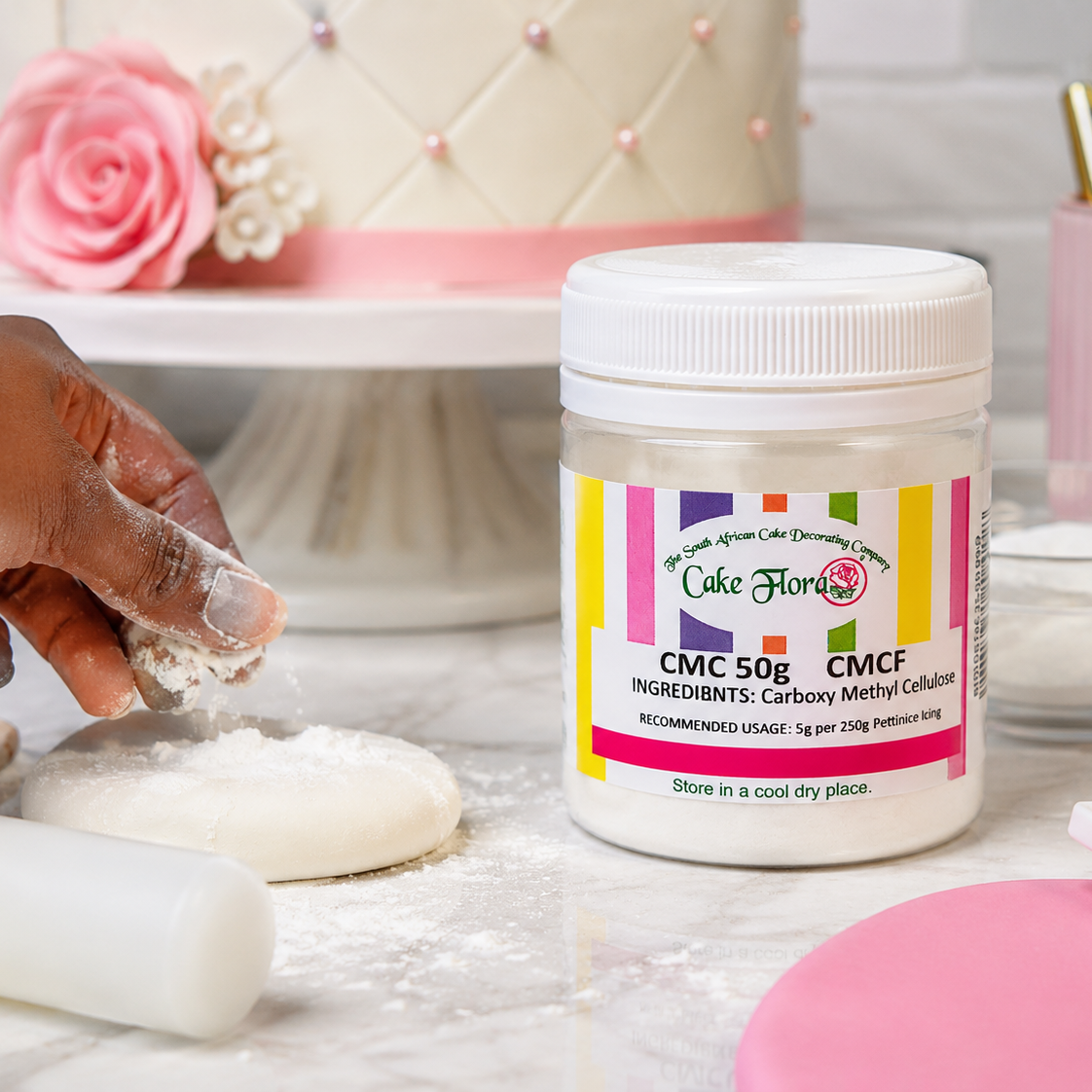 A jar of Cake Flowers CMC 50g Carb-Methyl Calorifics (CMC/CMCF) baking powder sits on a floured surface next to a hand shaping dough, with a pink decorated cake in the background.