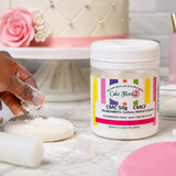 A jar of Cake Flowers CMC 50g Carb-Methyl Calorifics (CMC/CMCF) baking powder sits on a floured surface next to a hand shaping dough, with a pink decorated cake in the background.
