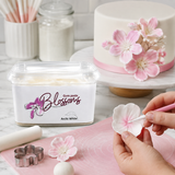 A tub of Arctic White Blossoms gum paste is shown in a kitchen setting next to a decorated cake, with hands working on a pink mat to shape a pink and white sugar flower.