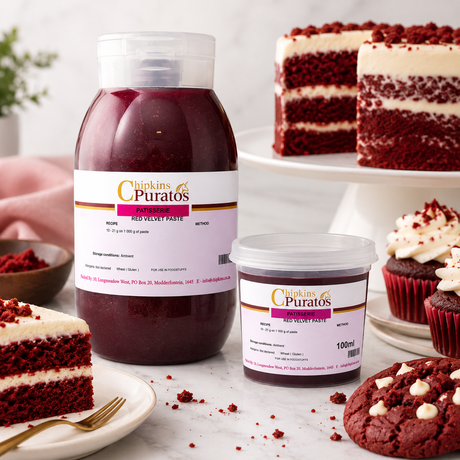 A large and a small jar of Chipkin's Puratos Red Velvet Paste surrounded by red velvet cake slices, cupcakes, and a chocolate cookie on a white marble surface.